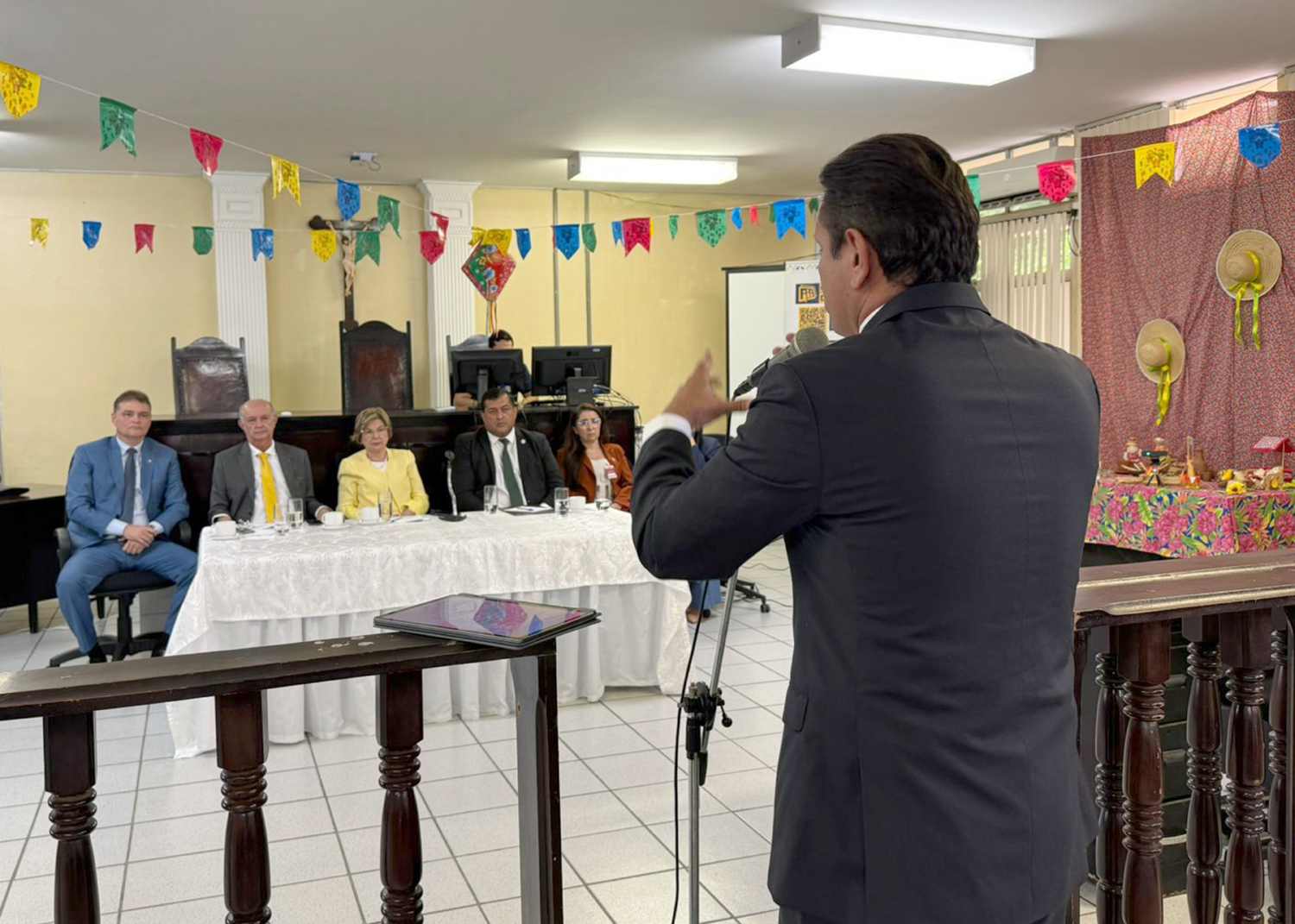 Fotografia colorida de um salão de audiências decorado com bandeirolas de festa junina. Em primeiro plano, um homem de terno, de costas para a câmera, fala ao microfone diante de uma mesa onde cinco autoridades estão sentadas. Ao fundo, aparecem a bancada do tribunal, um crucifixo na parede e computadores. À direita, uma decoração temática junina com chapéus de palha e tecidos coloridos. O ambiente é amplo e bem iluminado.