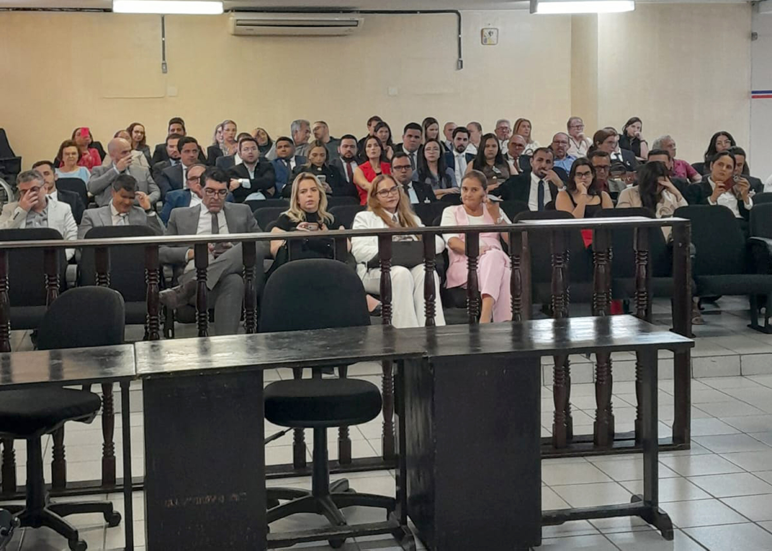 Fotografia colorida de um salão de audiências. Em primeiro plano, há mesas e cadeiras vazias. Ao fundo, dezenas de pessoas, entre magistrados, servidores e representantes de instituições, estão sentadas em fileiras, acompanhando atentamente a programação. O ambiente é amplo, climatizado, com piso claro, iluminação no teto e divisórias de madeira.