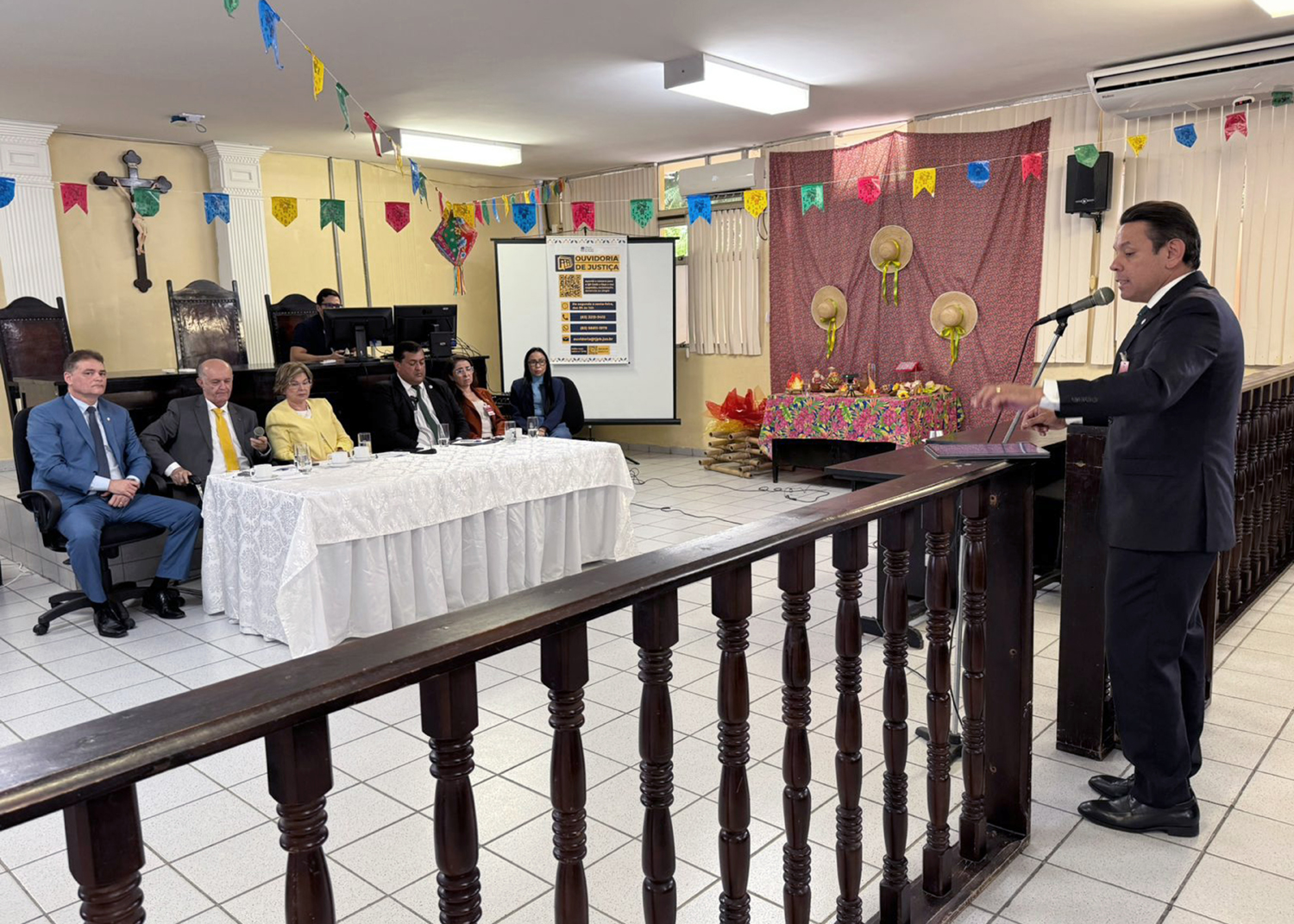 Fotografia colorida de um salão de audiências decorado com bandeirolas de festa junina. À direita, um homem de terno escuro fala ao microfone em um púlpito. À esquerda, cinco autoridades estão sentadas à mesa principal, coberta com toalha branca. Ao fundo, há um crucifixo na parede, um painel da Ouvidoria de Justiça e uma decoração temática com chapéus de palha e tecidos coloridos. O ambiente é amplo, iluminado e climatizado.